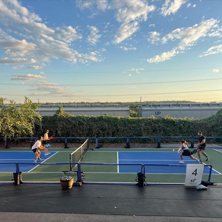 Four players in a doubles pickleball match on an outdoor blue-and-green court at golden hour, framed by hedges and freight train cars under a partly cloudy sky