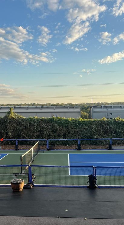 Four players in a doubles pickleball match on an outdoor blue-and-green court at golden hour, framed by hedges and freight train cars under a partly cloudy sky