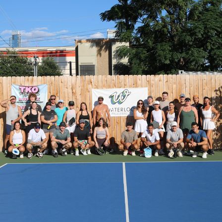 Large group of pickleball players smiling and posing on a blue outdoor court in front of a wooden fence with event banners, trees and city buildings in the background.
