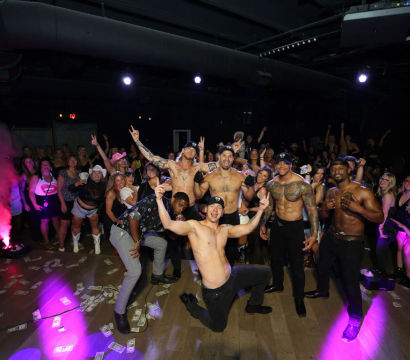 Shirtless male dancers strike poses on a nightclub stage in front of a cheering crowd, dollar bills scattered on the floor and colorful club lighting.