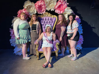 Five friends at a bachelorette party posing in an indoor event photo booth: the bride-to-be in a white dress, sash and cowboy hat sits on an ornate purple velvet and gold throne, surrounded by four smiling women in summer outfits in front of oversized pink and cream paper flowers.