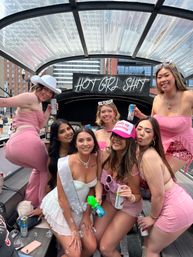 Seven women in pink and white outfits posing on a covered party boat in the city — bachelorette sash, bubble gun, canned drinks, and a neon 'HOT GIRL SH*T' sign overhead.