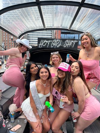 Seven women in pink and white outfits posing on a covered party boat in the city — bachelorette sash, bubble gun, canned drinks, and a neon 'HOT GIRL SH*T' sign overhead.