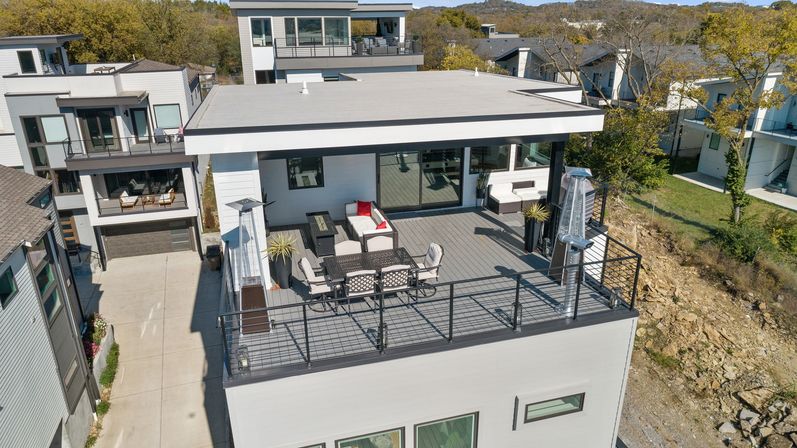 Modern rooftop deck on a multi-story home with dining table, cushioned lounge seating, tall patio heaters and metal railing overlooking neighboring contemporary houses and a wooded hillside.