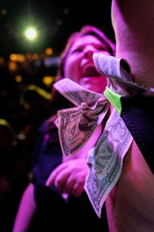 Close-up in a pink-lit nightclub: US dollar bills tucked into a performer’s costume at the arm, with a blurred cheering crowd and bright stage light in the background.