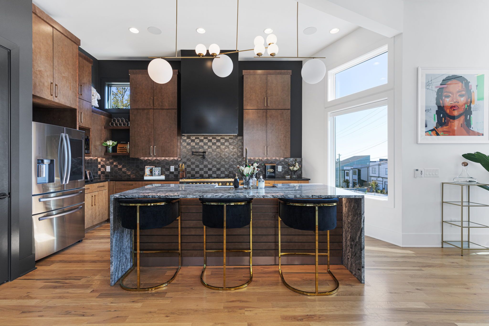 Stylish modern kitchen featuring a marble island with three gold-accented velvet bar stools, dark wood cabinets, stainless steel refrigerator, geometric tile backsplash, globe pendant lights, hardwood floors and large windows overlooking a neighborhood.