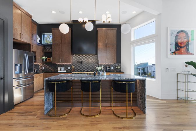 Stylish modern kitchen featuring a marble island with three gold-accented velvet bar stools, dark wood cabinets, stainless steel refrigerator, geometric tile backsplash, globe pendant lights, hardwood floors and large windows overlooking a neighborhood.