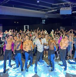 Six shirtless male dancers pose on a nightclub stage in front of a cheering crowd of women, dollar bills scattered on the floor and colorful stage lights creating an energetic party atmosphere.
