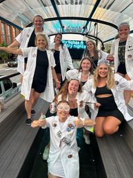 Group of women in lab coats and goggles celebrating a science-themed bachelorette party on an urban rooftop patio under a clear canopy