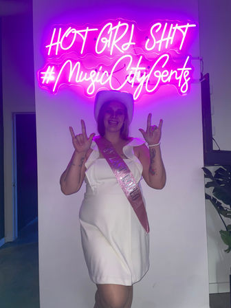 Person in a white dress and cowboy hat wearing a pink sash posing with rock-on hand signs under a bright pink neon sign reading “HOT GIRL SHIT #MusicCityGents” — playful Nashville party scene.