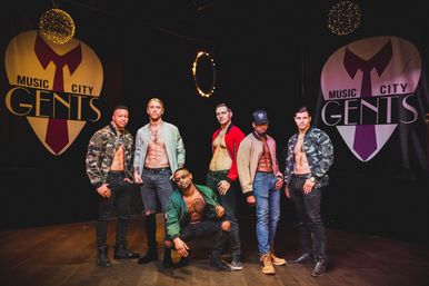 Six shirtless male dancers in bomber jackets and jeans posing under ring lights on a dim performance stage with large stylized heart banners — upbeat Nashville male revue vibe.