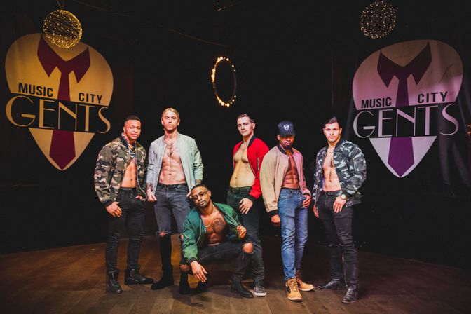 Six shirtless male dancers in bomber jackets and jeans posing under ring lights on a dim performance stage with large stylized heart banners — upbeat Nashville male revue vibe.