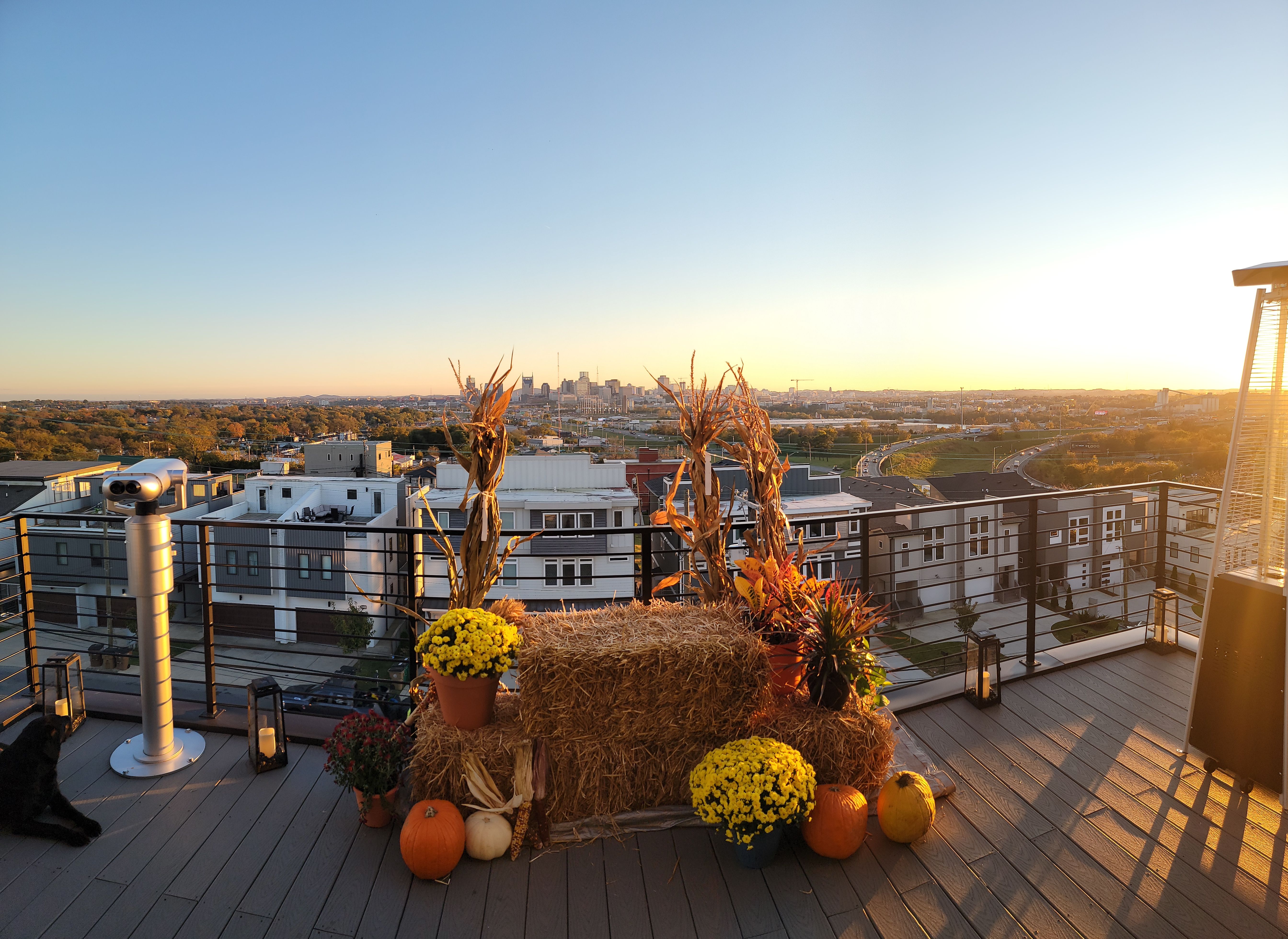 Rooftop terrace at golden sunset with fall harvest display — hay bales, pumpkins, potted mums and dried corn stalks beside a coin-operated viewer and patio heater, overlooking an urban skyline and winding highway.