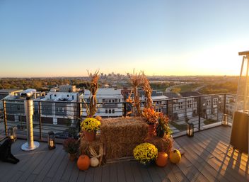 Rooftop terrace at golden sunset with fall harvest display — hay bales, pumpkins, potted mums and dried corn stalks beside a coin-operated viewer and patio heater, overlooking an urban skyline and winding highway.
