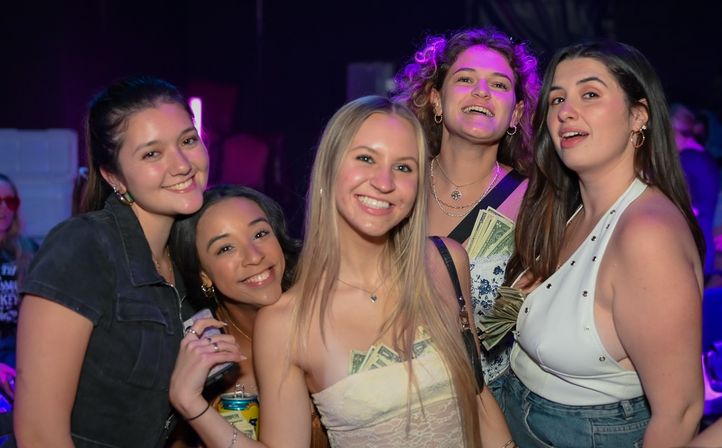 Five smiling women posing at a lively nightclub with purple lighting, some with cash tucked into their tops and one holding a canned drink — girls' night out urban nightlife scene.