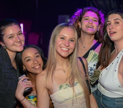 Five smiling women posing at a lively nightclub with purple lighting, some with cash tucked into their tops and one holding a canned drink — girls' night out urban nightlife scene.