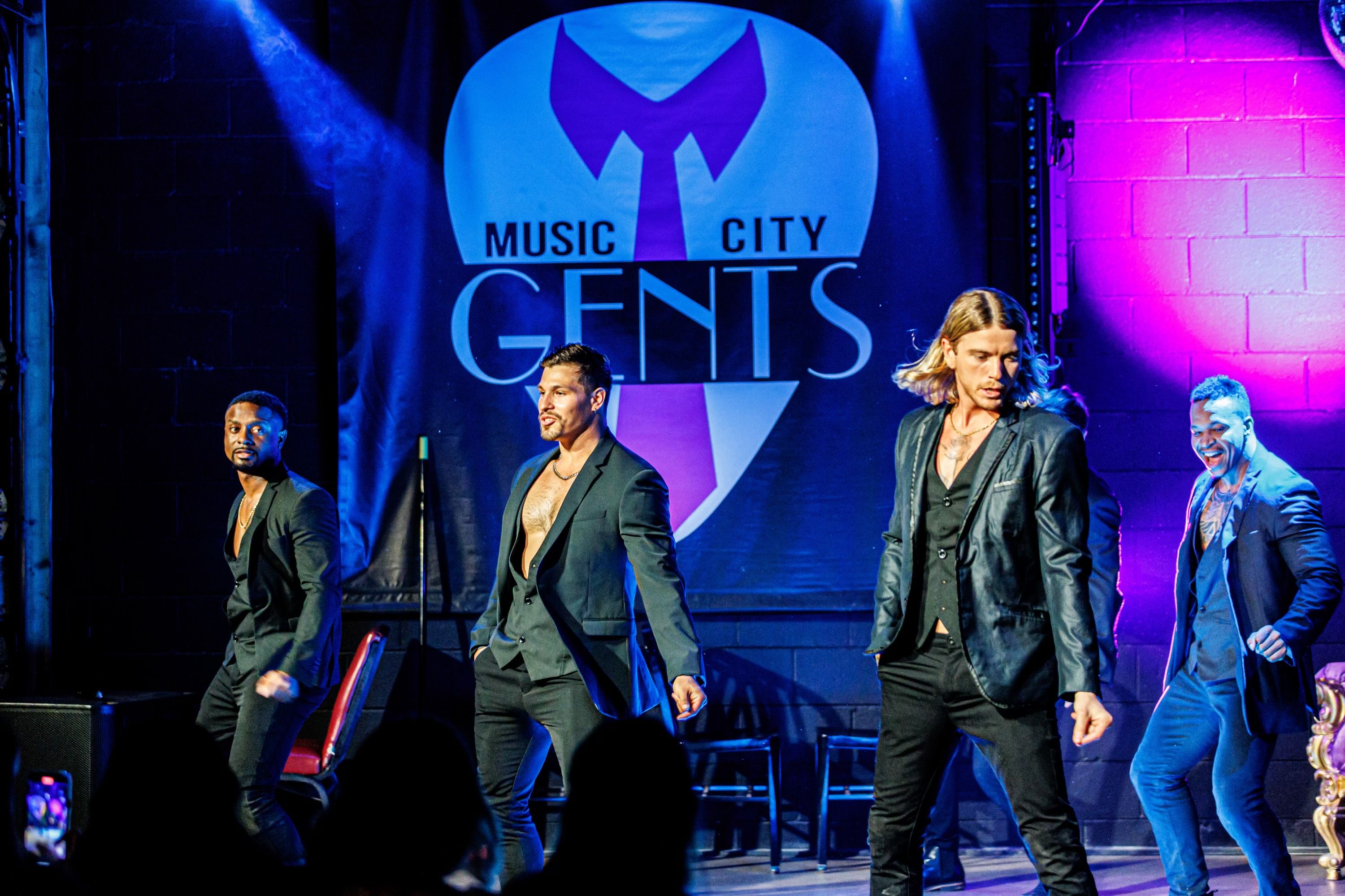Four charismatic male dancers in open-collar black suits performing on a neon-lit club stage with blue and purple spotlights and audience silhouettes in the foreground.
