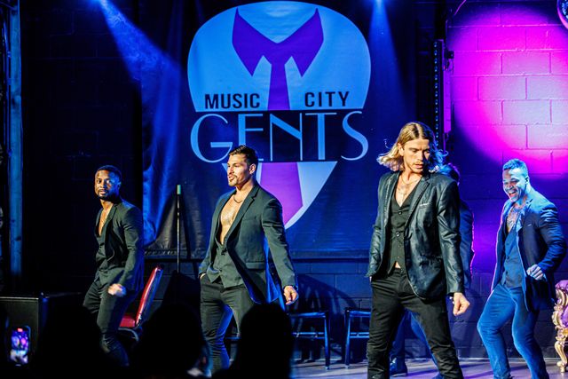 Four charismatic male dancers in open-collar black suits performing on a neon-lit club stage with blue and purple spotlights and audience silhouettes in the foreground.