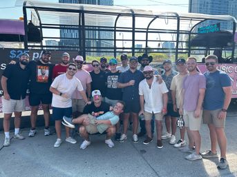 Group of about 17 men in casual summer clothes posing and smiling in front of a pink open-air party trolley with a city skyline behind them; some wear cowboy and pink hats and sunglasses while one man is playfully held horizontally in front.