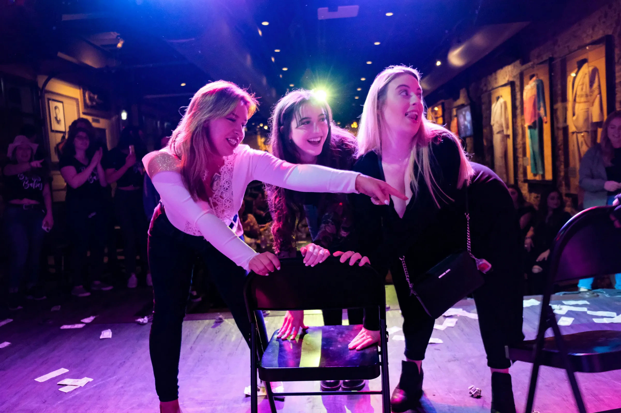 Three women laughing and leaning on a folding chair under purple stage lights at a packed live-music bar, pointing toward the crowd during a lively girls' night out.