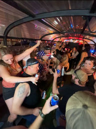 Crowded nighttime party bus with friends dancing and raising beer cans under colorful lights, including a rider wearing an American-flag outfit.