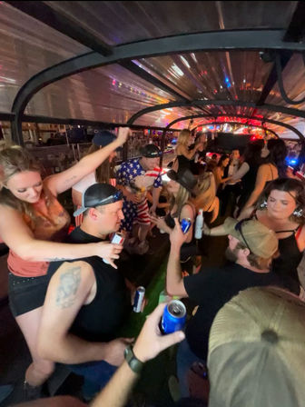 Crowded nighttime party bus with friends dancing and raising beer cans under colorful lights, including a rider wearing an American-flag outfit.