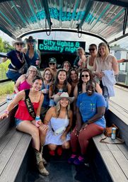Group of friends on a covered party wagon with a neon “City Lights & Country nights” sign, wearing cowboy hats and boots and holding canned drinks