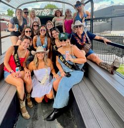 Bachelorette party group of women on a covered outdoor party wagon in a city setting, wearing cowboy hats, boots and sashes, smiling and holding drinks.