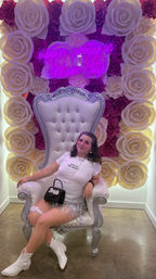 Person wearing a 'Wife of the Party' tee, silver fringe skirt and white boots sitting on a tufted white throne in front of an illuminated paper-rose wall with a pink neon sign reading 'sippin pretty in Music City' — playful Nashville bachelorette photo backdrop.