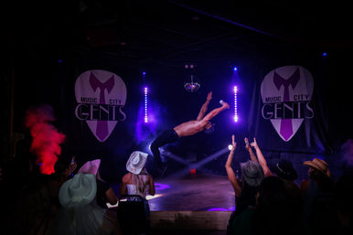 High-energy nightclub scene with a shirtless performer mid-backflip under purple spotlights and a disco ball, fog effects and LED columns as a cheering seated audience in cowboy hats reaches toward the stage.