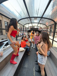 Group of friends partying on a covered open-air trolley rolling through a downtown city street, wearing summer outfits and boots, laughing, dancing and holding drinks under a clear arched canopy with city buildings visible