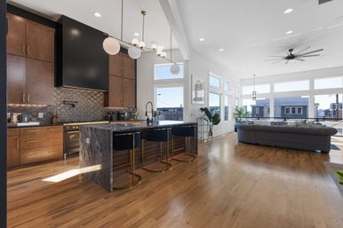 Modern open-concept kitchen and living room with marble island and brass-accent bar stools, wood cabinets, geometric tile backsplash, wide-plank hardwood floors, high ceilings, ceiling fan and large windows showcasing a city view.