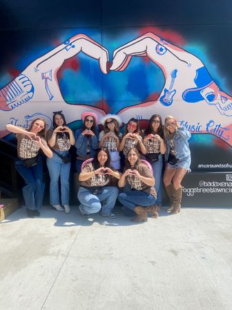 Group of friends in matching tees making heart shapes in front of colorful "Music City" hand-heart mural — outdoor Nashville street-art backdrop, festive outing