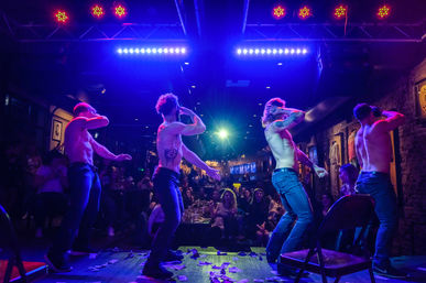 Four shirtless male dancers performing a choreographed strip show on a neon-lit nightclub stage with purple and blue spotlights, dollar bills on the floor and a cheering seated audience along exposed brick walls.