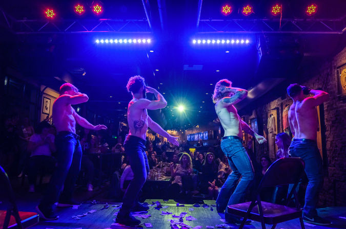 Four shirtless male dancers performing a choreographed strip show on a neon-lit nightclub stage with purple and blue spotlights, dollar bills on the floor and a cheering seated audience along exposed brick walls.