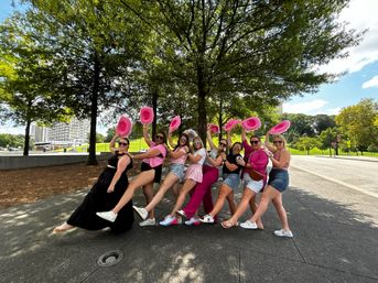 Eight friends in bright casual outfits and sunglasses strike a playful line under trees, each waving a pink cowboy hat in a sunny tree-lined urban plaza with buildings in the background.
