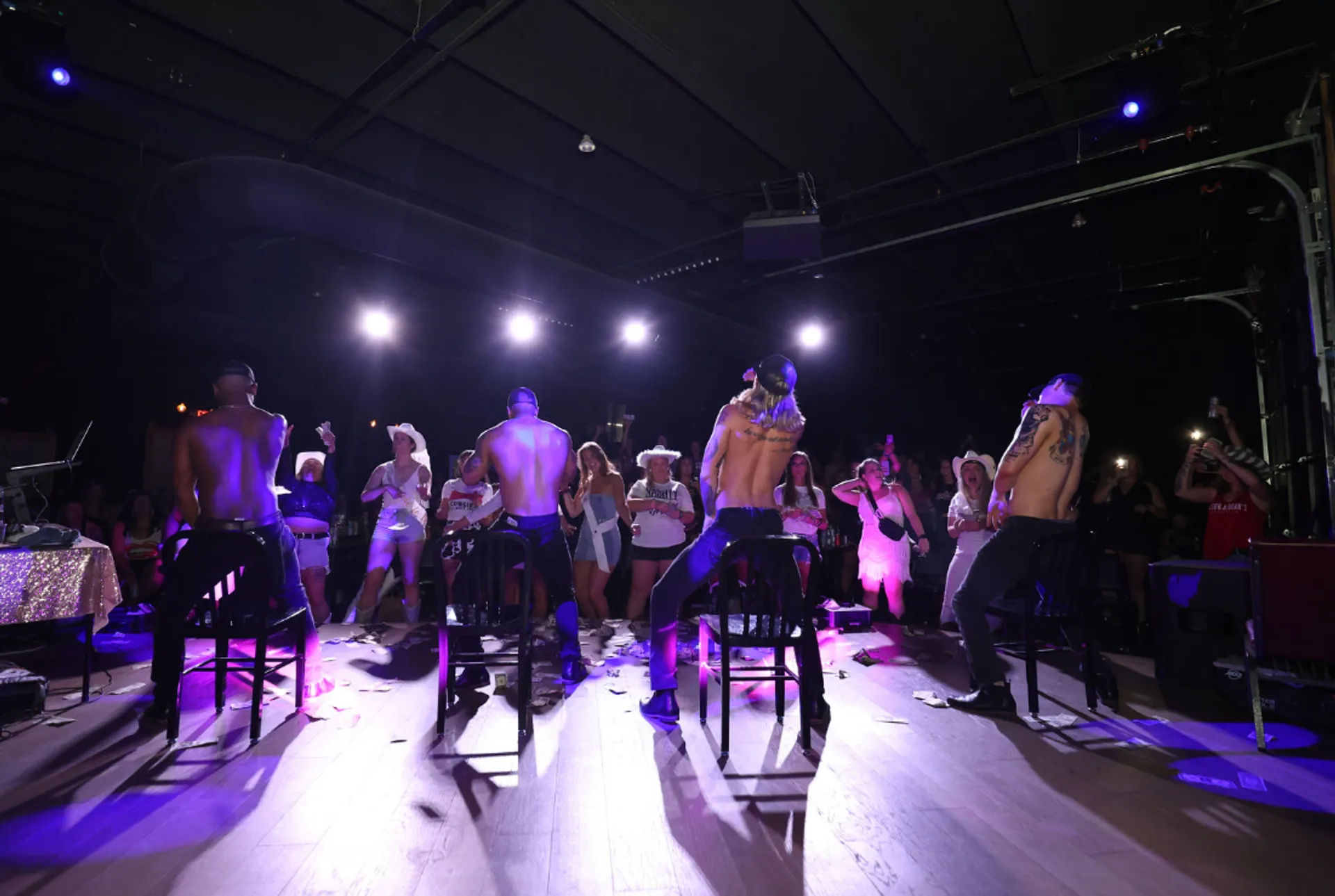 Four shirtless male dancers on stools performing under purple stage lights for a cheering crowd of women in a dim indoor club, with confetti on the floor and audience members filming.