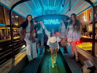 Five friends smiling inside a neon-lit party pedicab on a bustling downtown street at night, posing under a glowing neon sign for a night out.