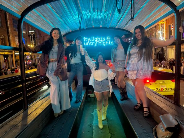 Five friends smiling inside a neon-lit party pedicab on a bustling downtown street at night, posing under a glowing neon sign for a night out.