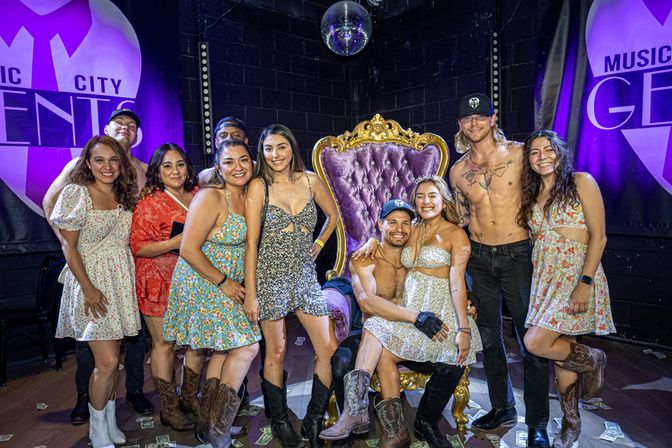 Group of friends posing on a music venue stage under purple lighting—women in summer dresses and cowboy boots gathered around an ornate purple-and-gold throne with two shirtless male performers, a disco ball overhead and dollar bills scattered on the floor.