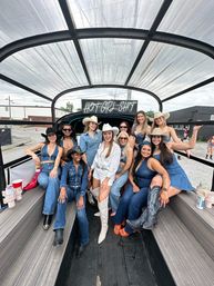 Group of women in denim and cowgirl hats smiling and posing inside a covered party trailer under a sign reading "HOT GIRL SHIT", wearing boots and western outfits with an urban street backdrop.