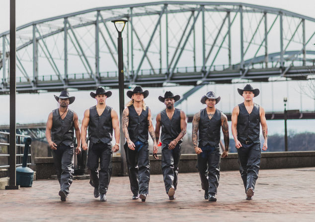 Six muscular men in black cowboy hats, leather vests and pants stride confidently along an urban riverfront promenade with a steel truss bridge overhead