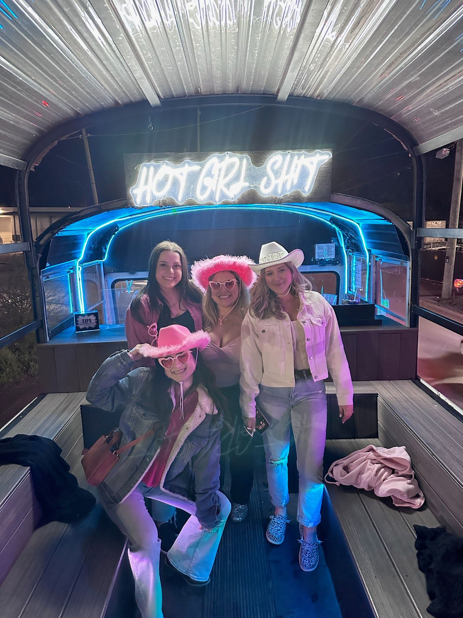 Four women wearing pink and white cowgirl hats posing inside a neon-lit party trailer with blue LED accents and a glowing 'HOT GRL SHXT' sign for a fun night out.
