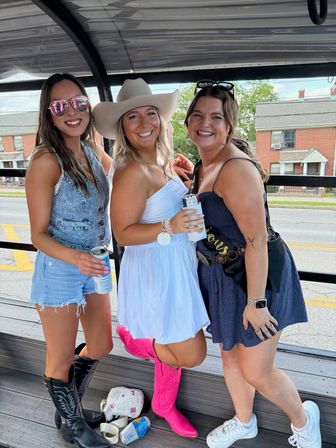 Three smiling friends on an open-air party trolley along a suburban street, wearing summer outfits — denim shorts with black cowboy boots, a white sundress with a cowboy hat and bright pink boots, and a navy dress with a sash — holding canned drinks and posing for a playful outing.