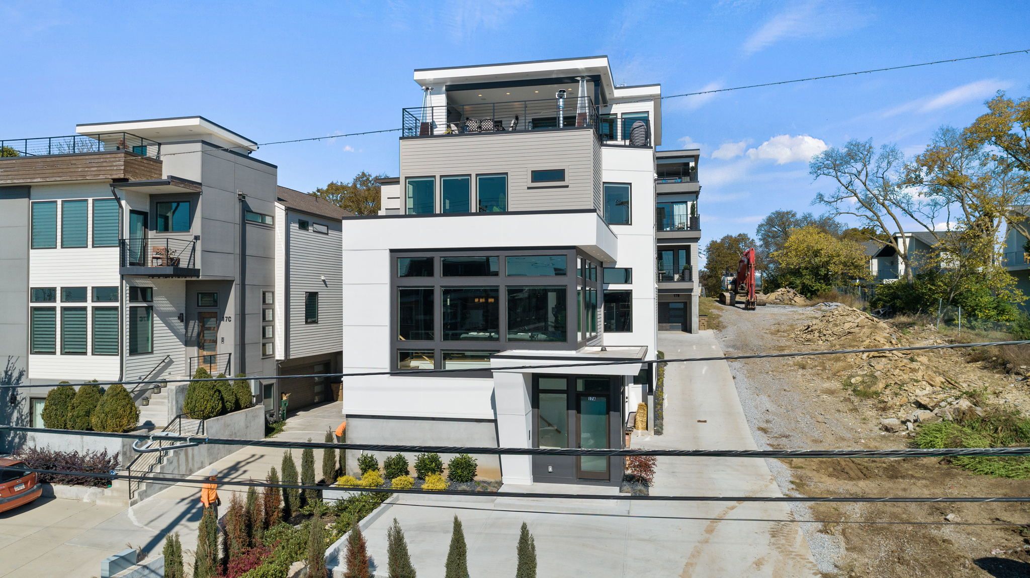 Contemporary multi-story white and gray townhouse with large glass windows and rooftop terrace, landscaped front walkway and concrete driveway, flanked by similar modern homes and a construction site under a clear blue sky.