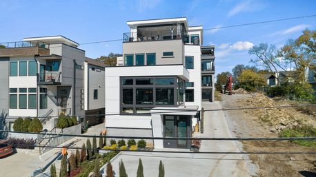 Contemporary multi-story white and gray townhouse with large glass windows and rooftop terrace, landscaped front walkway and concrete driveway, flanked by similar modern homes and a construction site under a clear blue sky.