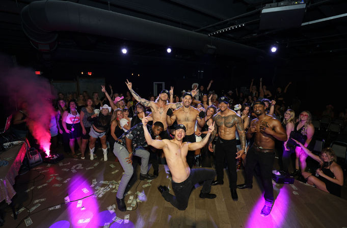 Shirtless male dancers posing on a nightclub stage with scattered dollar bills and colorful lights, surrounded by a cheering crowd enjoying a lively night out.