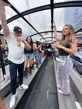 Group of adults cheering with drinks on a covered open-air party trolley rolling through a downtown street, casual outfits and cowboy boots, upbeat daytime bar-crawl vibe.