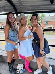 Three friends smiling on an open-air party trolley along a neighborhood street — center in a white dress and cowboy hat with bright pink cowgirl boots, left in denim shorts and black western boots, right in a navy dress and sneakers, all holding drinks.