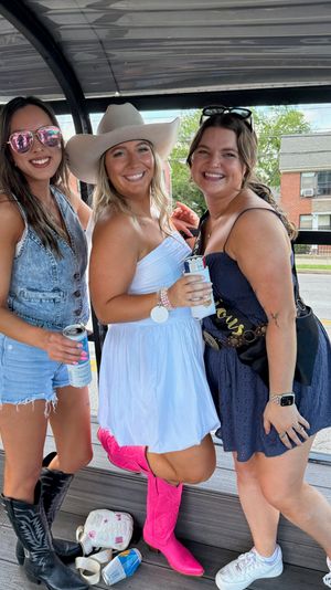 Three friends smiling on an open-air party trolley along a neighborhood street — center in a white dress and cowboy hat with bright pink cowgirl boots, left in denim shorts and black western boots, right in a navy dress and sneakers, all holding drinks.